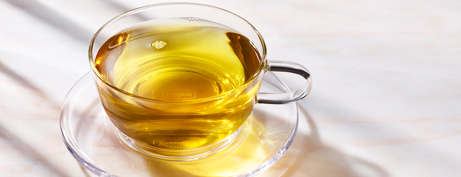 A transparent glass cup filled with golden green tea on a matching glass saucer