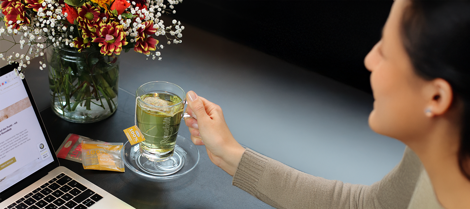 Frau sitzt am Tisch mit Laptop, hält eine Tasse Tee von Ronnefeldt in der Hand; daneben ein Blumenstrauß in einem Glas und mehrere Teeverpackungen.