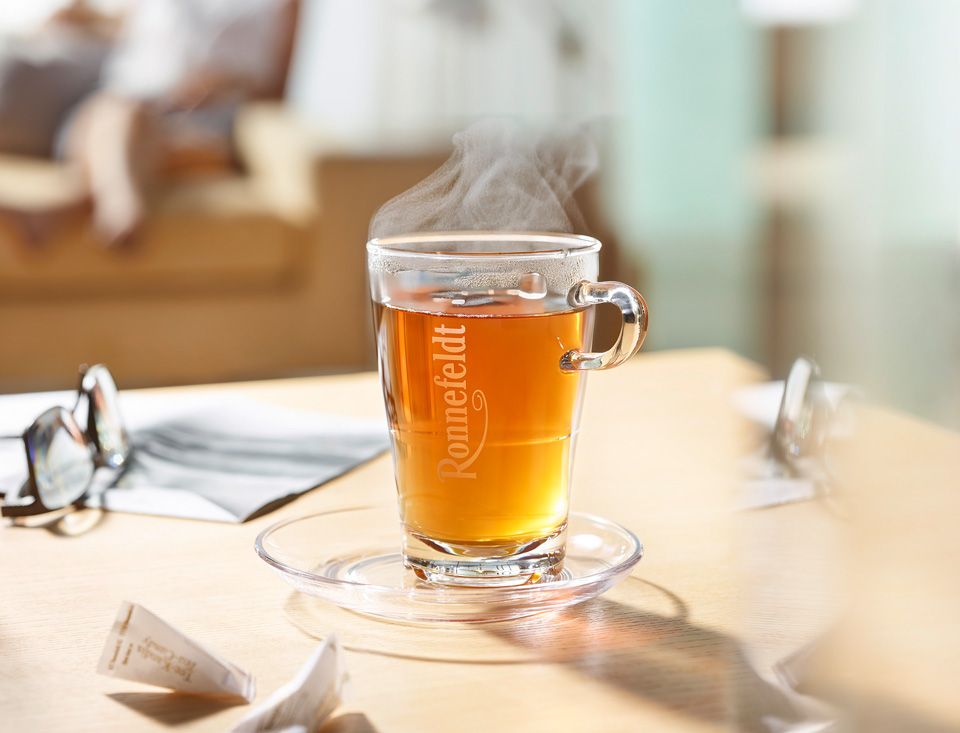 Ronnefeldt Teetasse mit braunem Tee und im Hintergrund eine Frau auf einer Couch