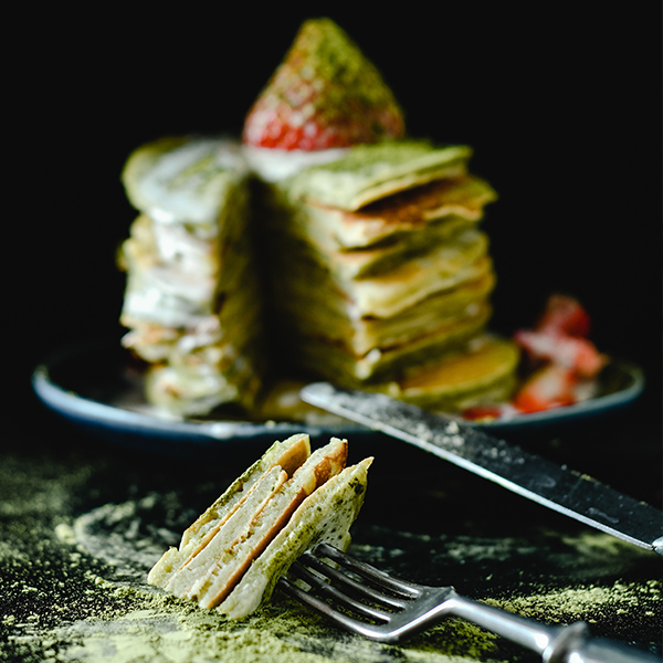 Matcha-Pancakes mit Erdbeere, im Anschnitt präsentiert, mit einem Stück auf einer Gabel im Vordergrund und Matcha-Pulver auf dunklem Hintergrund