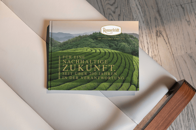 Ronnefeldt Nachhaltigkeitsbroschüre mit Teeplantagen-Motiv liegt auf einem Sessel.