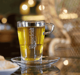 Glastasse mit Ronnefeldt Tee auf Unterteller vor warmem, unscharfem Hintergrund.