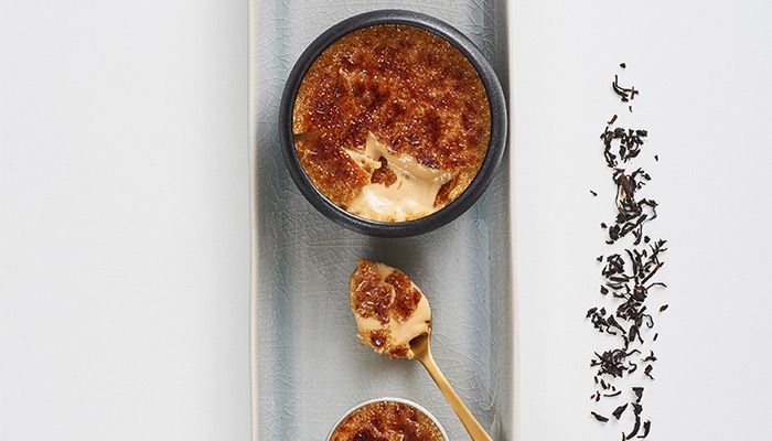 Crème brûlée mit karamellisierter Kruste, serviert mit goldener Löffelportion und losen Teeblättern als Dekoration