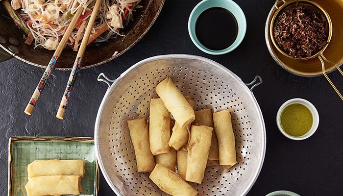 Asiatische Frühlingsrollen in einem Sieb mit verschiedenen Dips und Nudelgericht auf dunklem Untergrund