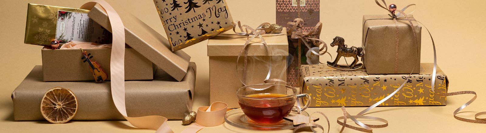 Weihnachtlich verpackte Geschenke, Schleifen und Deko-Elemente arrangiert um eine dampfende Tasse Tee auf beige-goldenem Hintergrund.