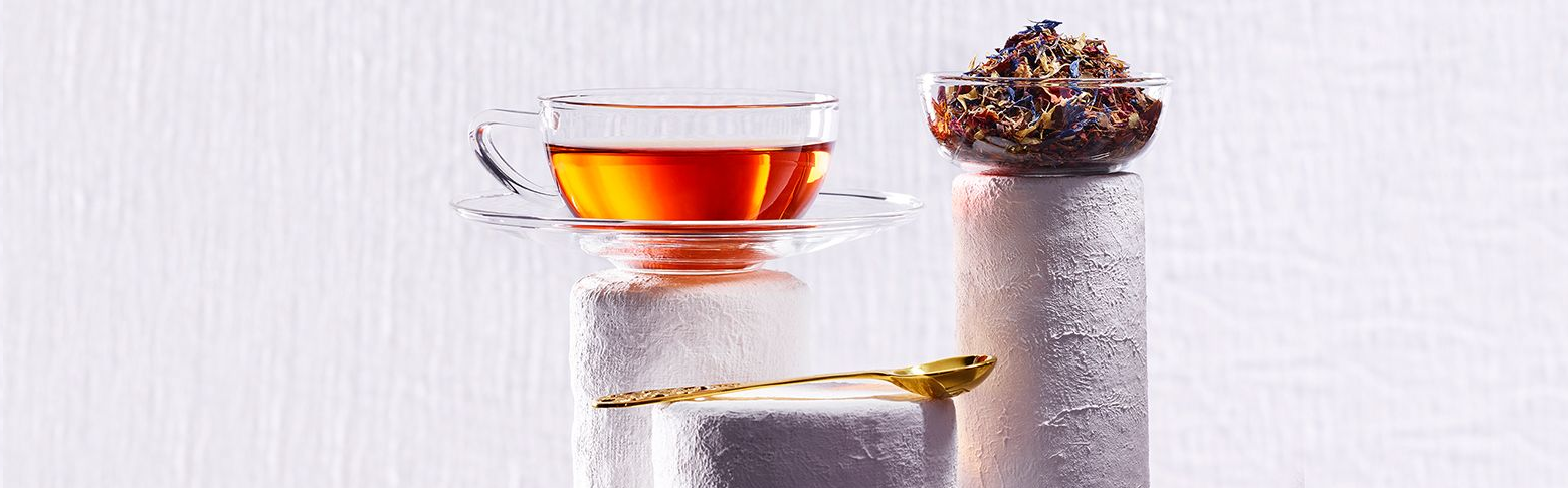 Glass cup of amber-colored tea, bowl of colorful loose tea, and a golden teaspoon on white textured pedestals.