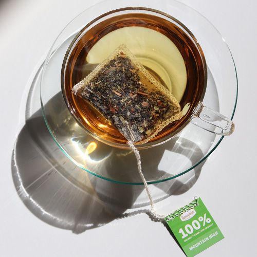 Glastasse mit Kräutertee und einem durchsichtigen Teebeutel auf einer Glas-Untertasse mit Lichteinfall und Schattenwurf.