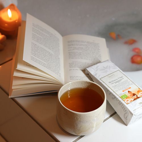 Ronnefeldt Wellness Chai Tee mit einer Teetasse Tee, einem Buch und Kerzenlicht, im Hintergrund ein Schaumbad mit Rosenblüten.