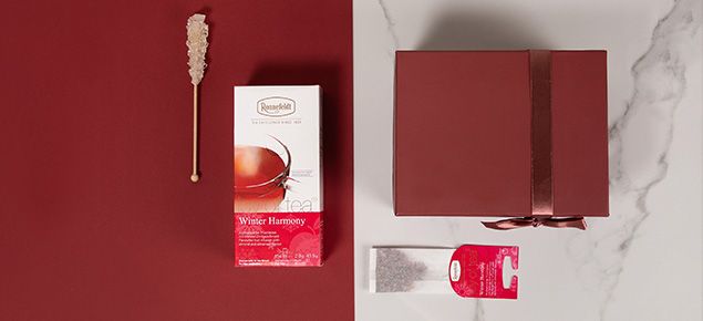 Teebox mit Teekandis-Stäbchen, Teebeutel und Geschenkbox liegen halb auf roten Untergrund und Marmortisch