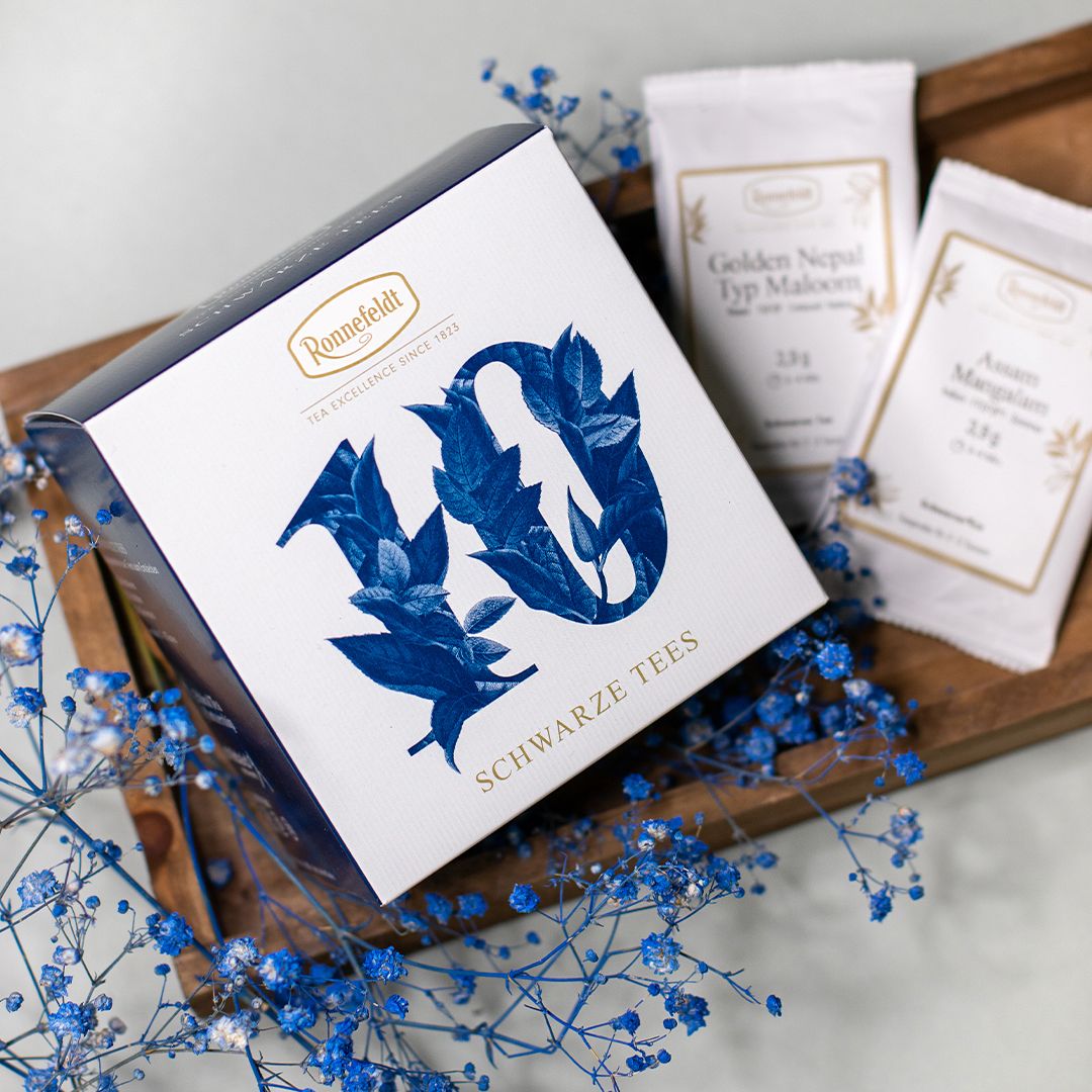 Ronnefeldt Probierbox - Schwarze Tees, dekoriert mit blauen Trockenblumen und edlen Einzelverpackungen im Hintergrund.