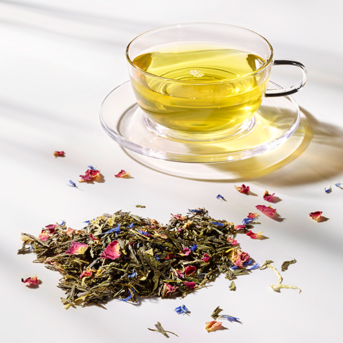 Glastasse mit goldenem Morgentau-Tee auf einer Glas-Untertasse, dekoriert mit losem Grüntee mit Rosenblüten und blauen Kornblumen.