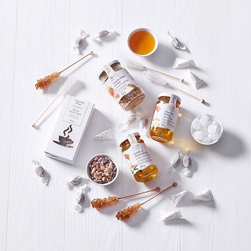 Ronnefeldt Süßigkeiten wie Honig, Kandis-Sticks, Teebonbons und ein Vanille-Sahne Zucker auf weißem Hintergrund, ergänzt durch eine Teetasse.