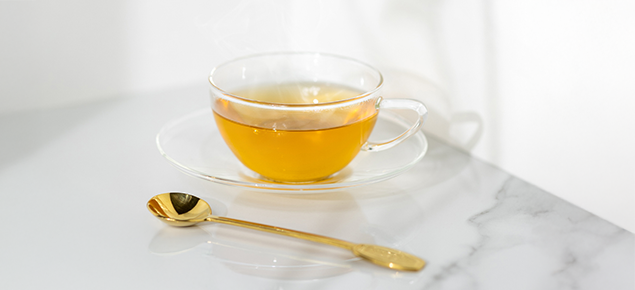 Eine Glasstasse mit goldenem Tee auf einer Marmoroberfläche, daneben ein goldener Teelöffel